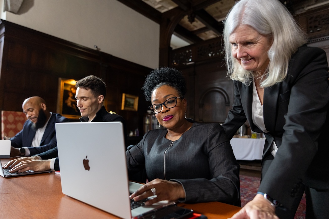 Two women in focus, one seated at laptop and the other looking at the screen over her shoulder, seated at board room table.