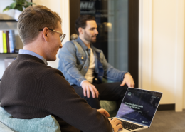 Two men are in a casual office setting, one sitting on a chair typing on a laptop, wearing glasses and a brown sweater. The other, wearing a denim jacket, is seated on a couch in the background.