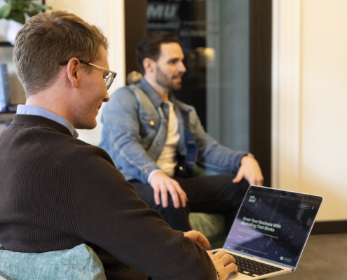Two men are in a casual office setting, one sitting on a chair typing on a laptop, wearing glasses and a brown sweater. The other, wearing a denim jacket, is seated on a couch in the background.