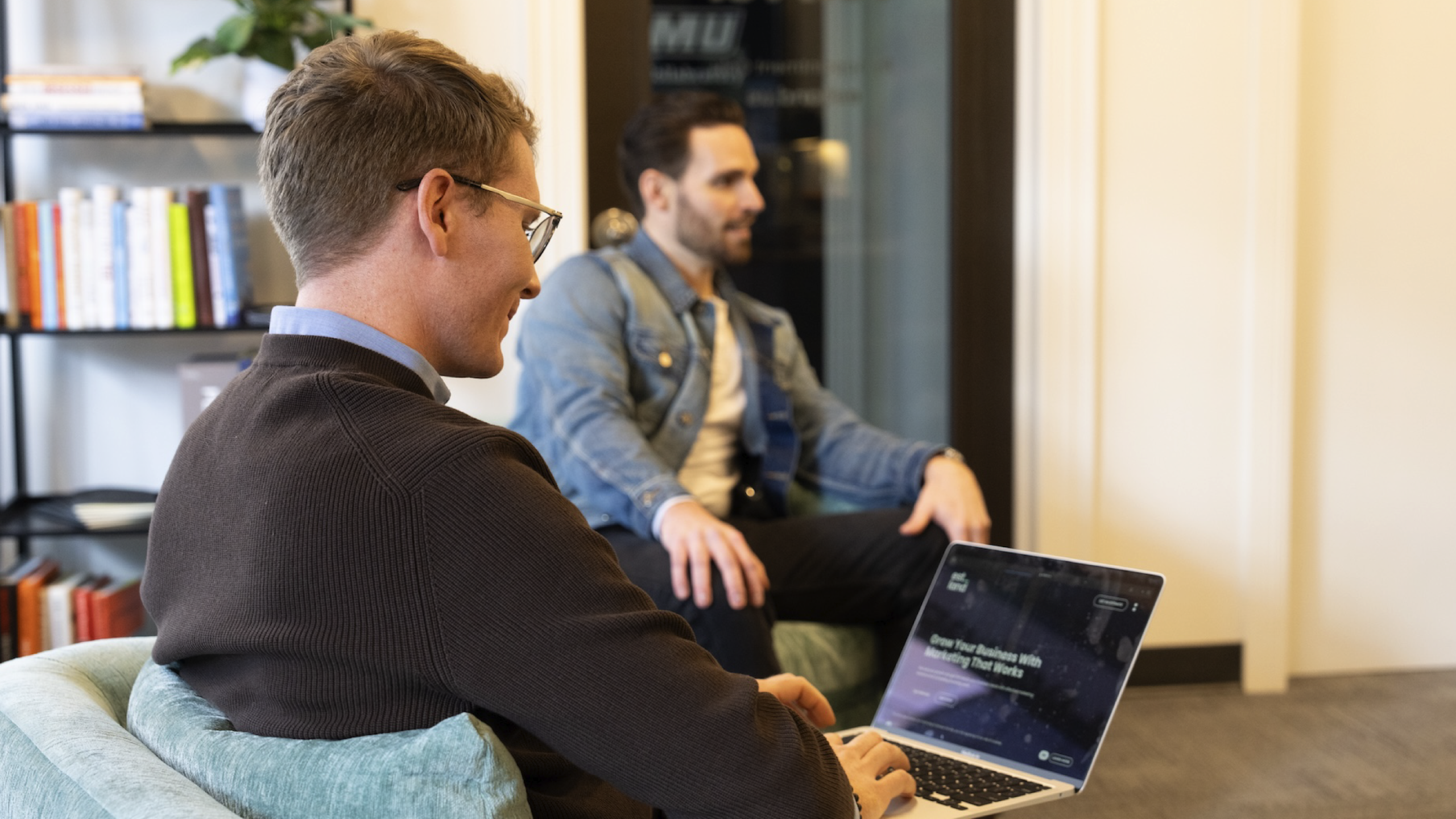 Two men are in a casual office setting, one sitting on a chair typing on a laptop, wearing glasses and a brown sweater. The other, wearing a denim jacket, is seated on a couch in the background.