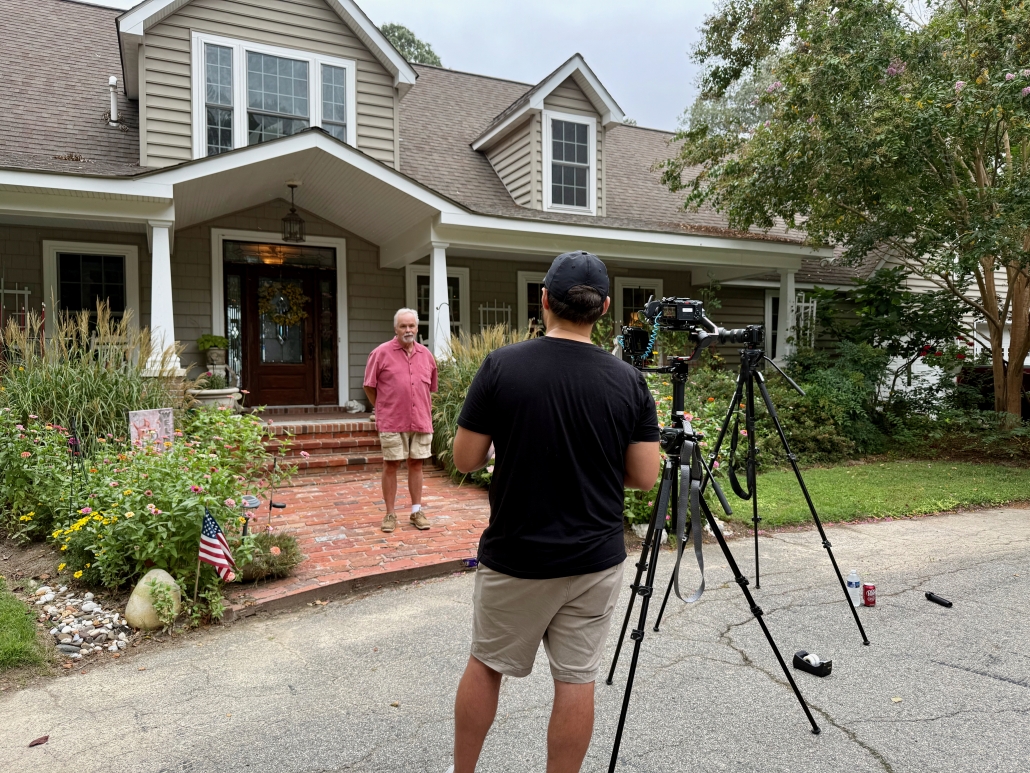 Behind-the-scenes of an Estland photo and video shoot. A person is being filmed outside a house, with trees and bushes in the background. The house has a porch and the man is in a casual conversation with Estland's videographer.