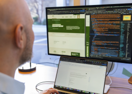 A person sits at a desk with a laptop, focusing on a large monitor displaying web development code and a website layout side by side.