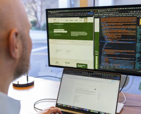 A person sits at a desk with a laptop, focusing on a large monitor displaying web development code and a website layout side by side.