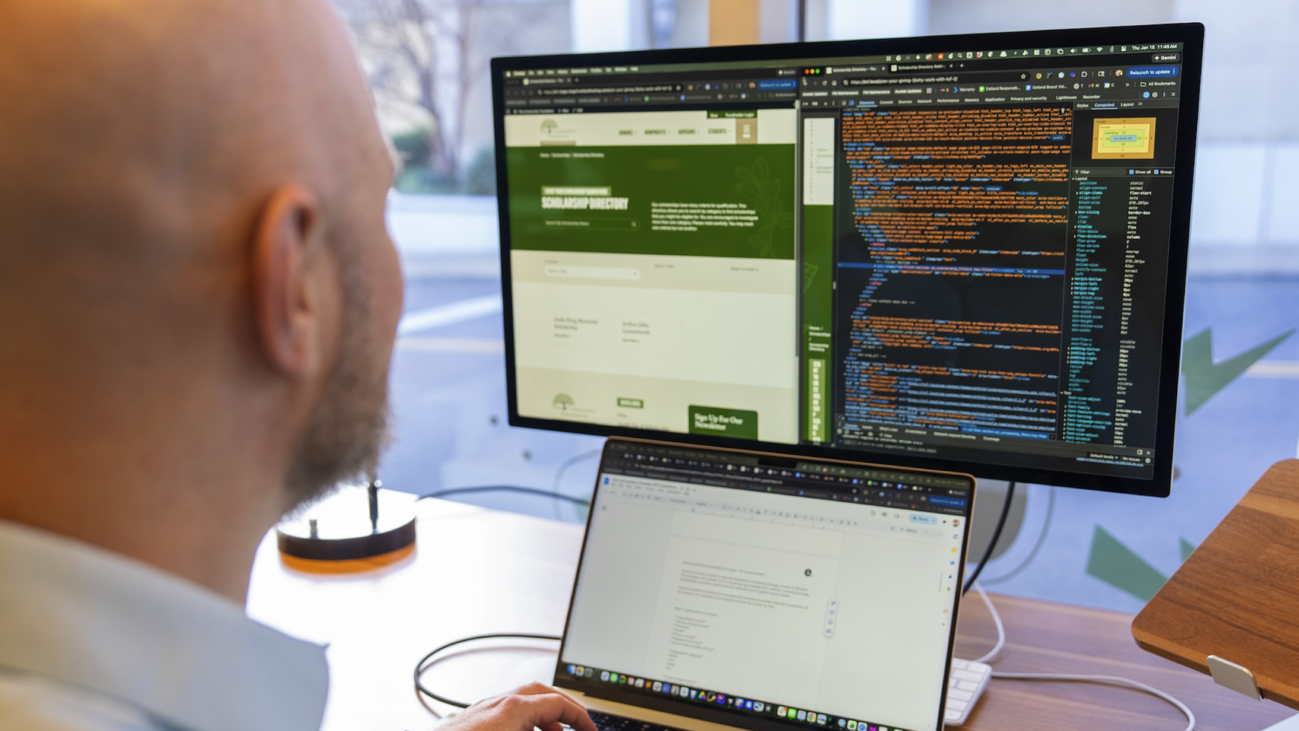 A person sits at a desk with a laptop, focusing on a large monitor displaying web development code and a website layout side by side.